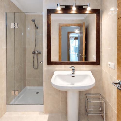 Baño moderno en apartamento de Baiona con lavamanos pedestal, espejo enmarcado, ducha de cristal y paredes con azulejos beige