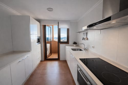 Cocina blanca moderna con vistas al mar, electrodomésticos de acero inoxidable y acceso a balcón en apartamento de Baiona