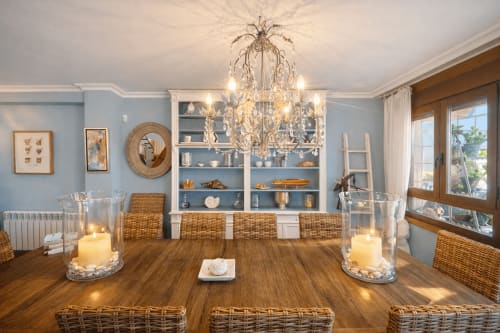 Elegante comedor del apartamento de Baiona con mesa de madera, sillas de mimbre, lámpara de cristal y vistas al mar