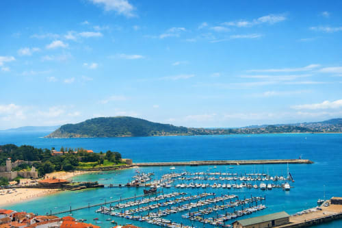 Puerto costero de Baiona con embarcaciones amarradas, mar azul, península arbolada y edificios de pueblo con tejados rojos