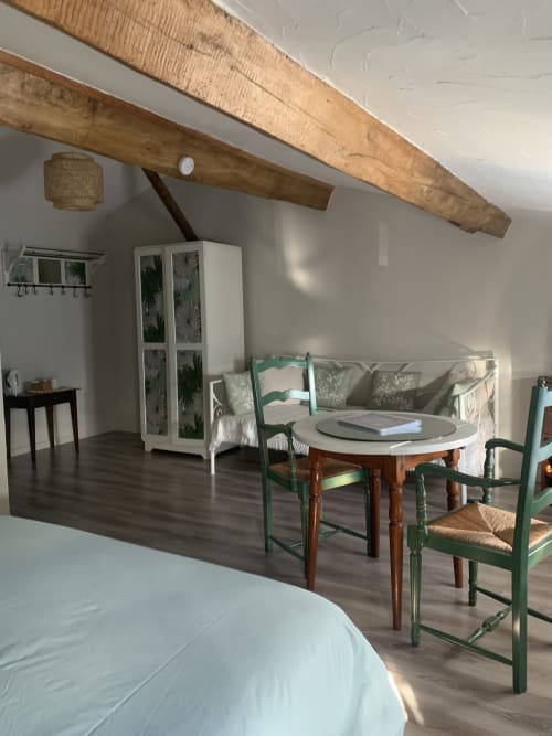 Le coin salon de la Chambre du Marais, comprenant une table ronde, deux chaises en bois vert, un canapé en fer forgé blanc et une armoire customisée sous de grandes poutres apparentes.