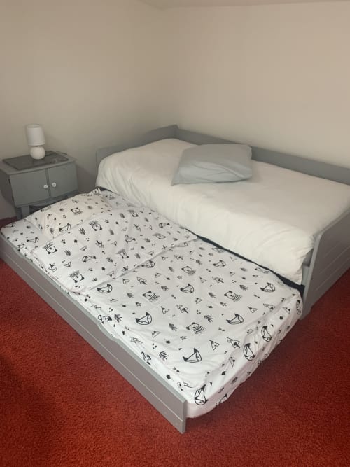 Le lit gigogne de la Chambre Familiale Soleil déplié pour offrir deux couchages enfants, posé sur une moquette rouge avec une table de chevet grise assortie.
