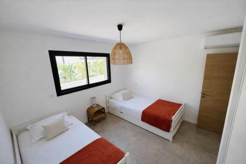 Bright twin bedroom in Villa Corail Favone, with woven pendant light and window view, managed by Corsu di Core