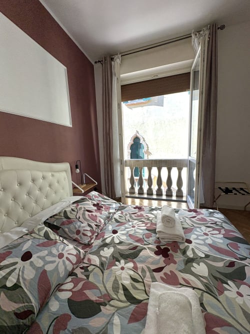 Camera da letto a Le Arche B&B a Verona con testata trapuntata color crema, biancheria floreale e balcone con vista sull'architettura cittadina