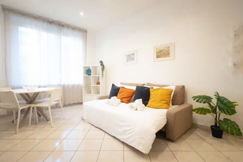 Moderno monolocale a IV House Roma con divano letto beige, biancheria bianca, cuscini gialli, zona pranzo e piante verdi