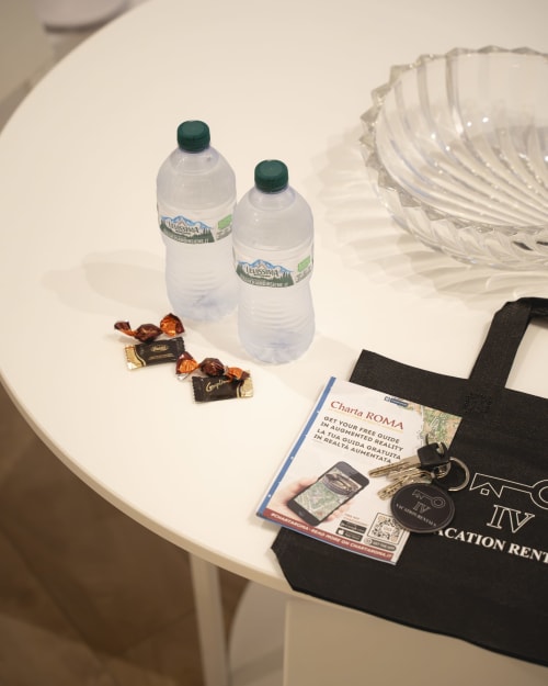 Amenità di benvenuto al tavolo di IV House Rome: bottiglie d'acqua, cioccolatini, chiave della camera, guida Charta Roma e bicchieri di plastica