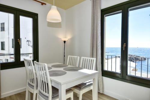 Bright seafront dining room with white table, chairs, pendant lights, and sea views through green-framed doors at Palafrugell