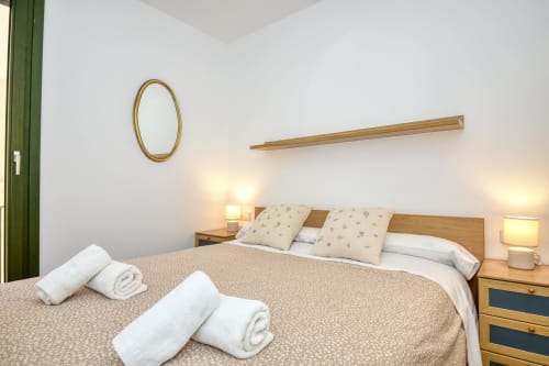 Cozy bedroom with wooden bed, pillows, rolled towels, bedside lamps, oval mirror, and green curtains in Palafrugell apartment