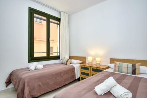 Bright seafront apartment bedroom in Palafrugell with two beds, green-framed window, wooden nightstands, and white linens