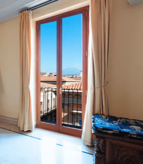 Camera moderna con porte-finestre che si aprono su balcone con vista sui tetti di Catania e le montagne dell'Aparthotel Bentivoglio