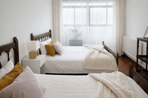 Dormitorio luminoso en apartamento Holibai Amieiro, Baiona con camas gemelas, marcos de madera, sábanas blancas, almohadas mostaza y vistas ciudad