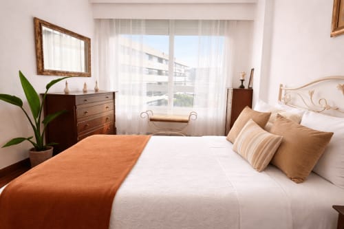 Dormitorio luminoso en apartamento de Baiona con vistas a la ciudad, muebles de madera, manta naranja, ropa de cama blanca y decoración contemporánea