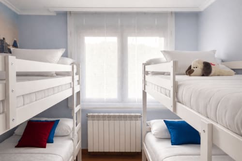 Dormitorio infantil luminoso con cuatro literas blancas, almohadas azules y rojas, peluches y grandes ventanas en Holibai