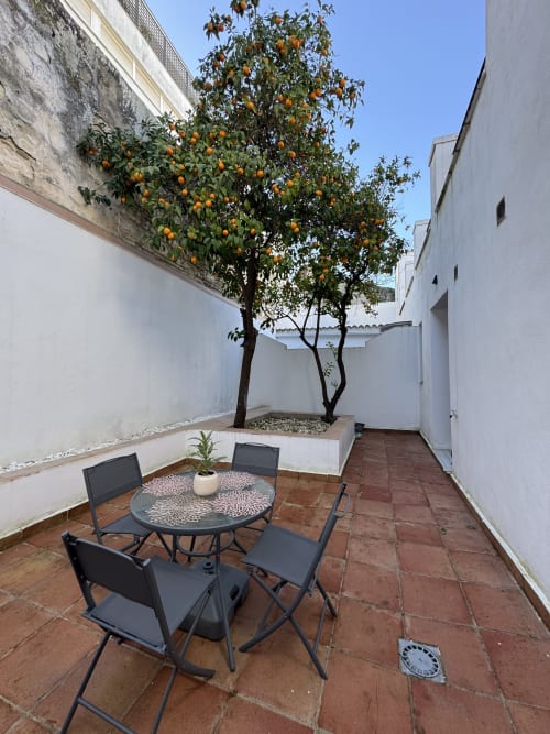 Patio con naranjo, mesa redonda, sillas negras y paredes blancas en apartamento Wehost Costaluz Puerto Santa María