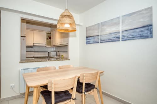 Modern dining area with wooden table and woven pendant light, open kitchen view, oceanview artwork in beachfront apartment.