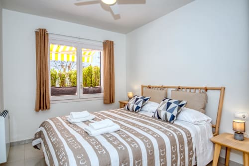 Bright beachfront bedroom in Palafrugell apartment with wooden bed, striped bedding, and sea view through large windows.