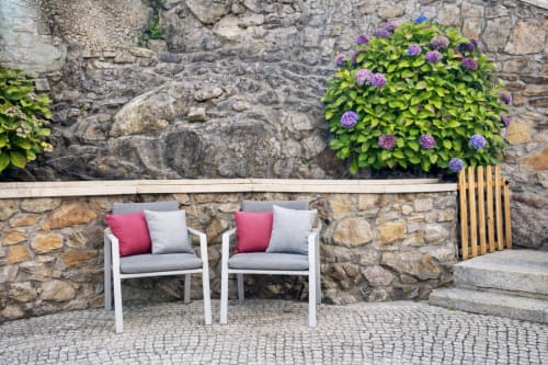 Dos modernas sillas de exterior blancas con cojines rosa en una terraza pavimentada de la villa Holibai en Baiona, respaldadas por un muro de piedra con