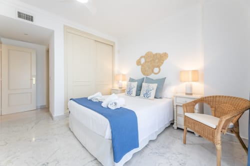 Bright modern bedroom in Marbella apartment with king bed, blue accents, wicker chair, and beige wall art