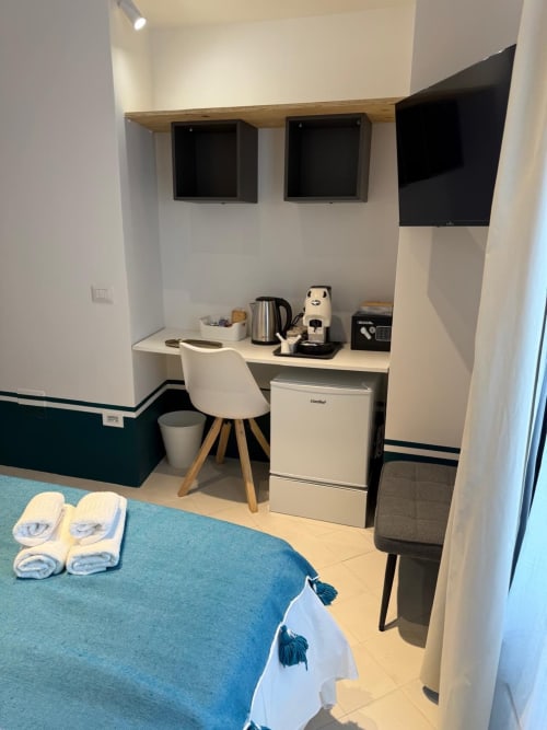 Angolo cucina compatto in hotel con scrivania, sedia bianca, frigorifero, bollitore elettrico, macchina da caffè e TV a parete