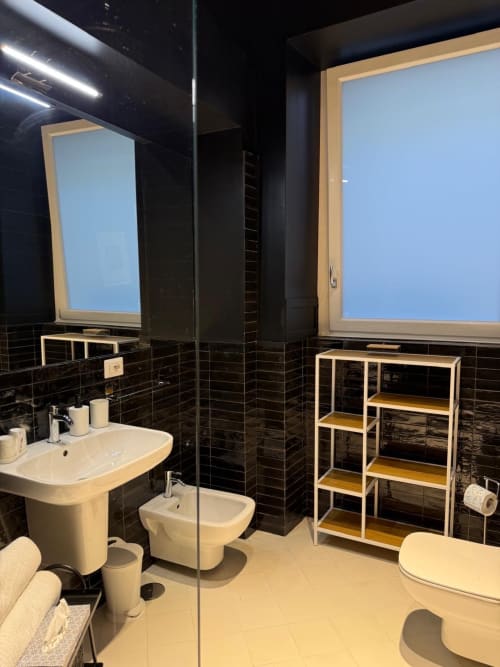 Bagno moderno nella suite Porta Capuana dell'hotel Tresette di Napoli, con rivestimenti in piastrelle nere, lavabo bianco, bidet e dettagli in legno