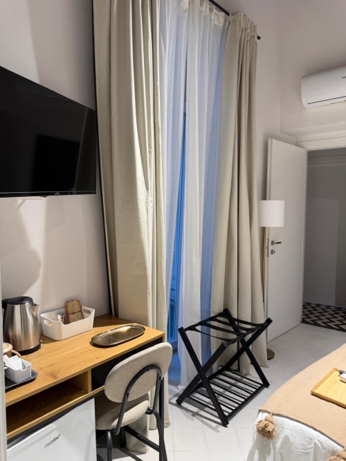 Camera moderna dell'hotel Tresette Porta Capuana a Napoli con scrivania in legno, tende beige, TV a schermo piatto e bagaglio nero