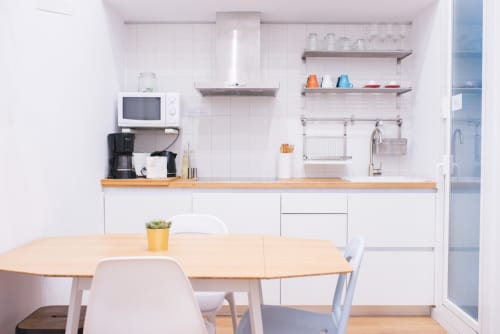 Moderna cocina de apartamento barcelonés con armarios blancos, encimera de madera, mesa de comedor y equipada con microondas y cafetera
