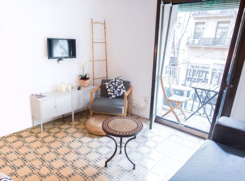 Sala de estar de apartamento moderno en Barcelona con vistas al balcón, amueblada con sillón gris, aparador blanco, TV y suelo geométrico