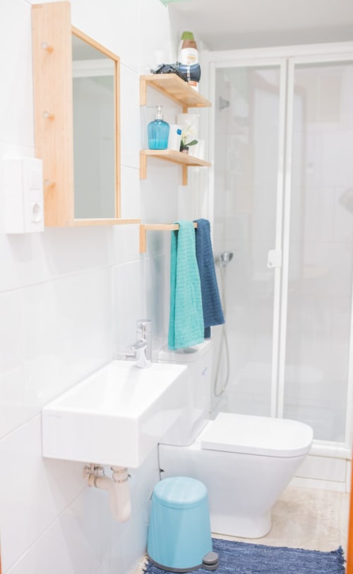 Baño moderno en apartamento de Barcelona con estantería de madera, lavabo blanco, inodoro, ducha y toallas turquesas