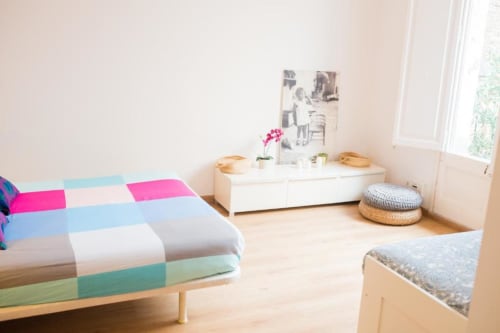 Luminoso dormitorio de apartamento barcelonés con ropa de cama colorida, cómoda blanca, suelos de madera y vistas a la ciudad desde la ventana