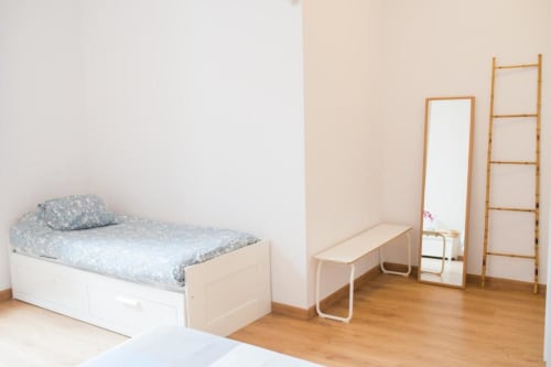 Luminoso dormitorio de apartamento barcelonés con cama individual, estantería de escalera de madera, espejo y banco en estudio minimalista