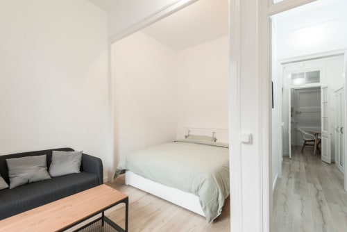 Dormitorio de apartamento moderno en Barcelona con sofá negro, cama blanca, suelos de madera y distribución abierta
