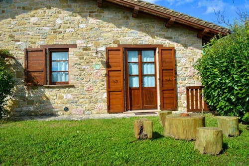 Stone cottage exterior at Agriturismo Colle Mura in Gualdo Tadino with wooden doors, windows, green lawn and tree stumps