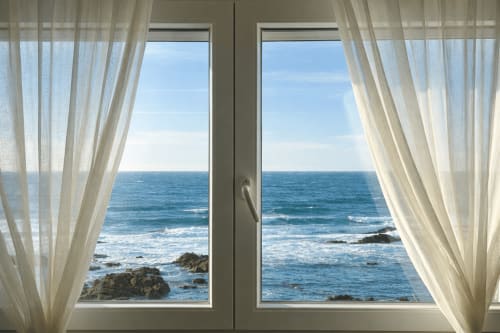 Cortinas translúcidas enmarcan puertas abiertas con vistas panorámicas de la costa de Baiona, rocas y olas desde el Albatros Dúplex