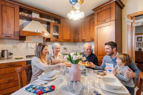 Famiglia multigenerazionale festeggia la Festa del Papà nella calorosa cucina in legno di Casa Meravigliosa, tra cibo e tulipani