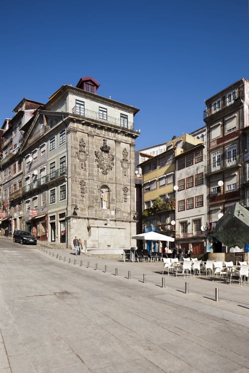 Praça histórica do Porto com edifícios coloridos de azulejo, mesas de café ao ar livre e arquitetura em pedra sob céu azul claro