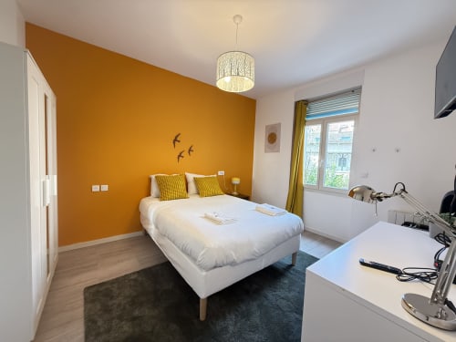 Chambre moderne dans un appartement de Metz avec un mur d'accent orange, lit blanc, coussins jaunes, suspension, bureau et vue sur la ville