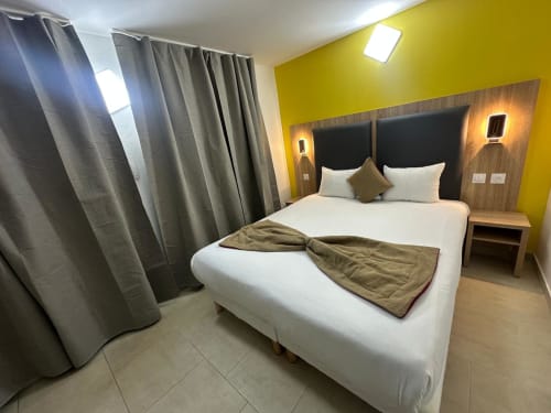 Modern double bedroom at Hôtel Norotel Brumath with white bed, yellow accent wall, wooden headboard, and contemporary