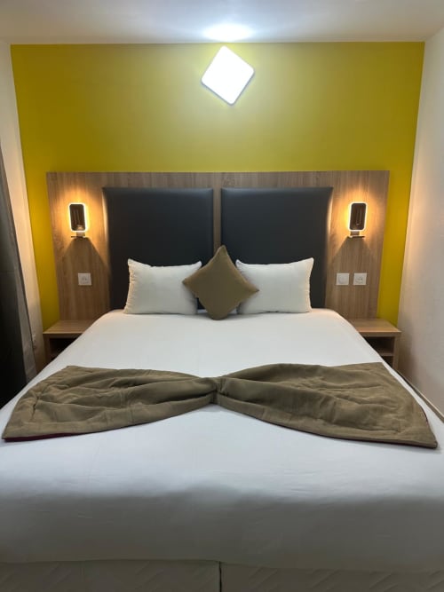 Modern double bedroom at Hôtel Norotel Brumath with yellow accent wall, dark headboards, and olive green throw pillows