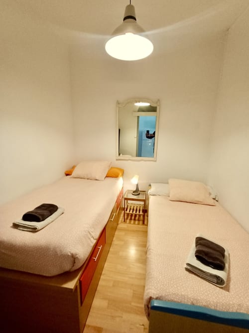 Compact twin bedroom in Barcelona apartment with two beds, pendant light, mirror, and wooden flooring