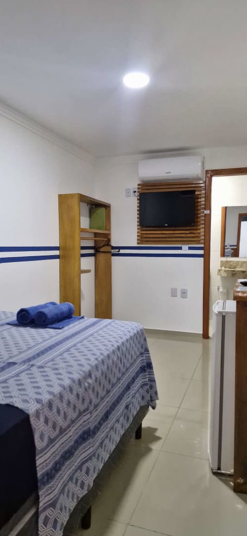 Compact private suite at Praieira Pousada in Itacaré with blue-striped bed, flat-screen TV, wooden furnishings, and AC unit