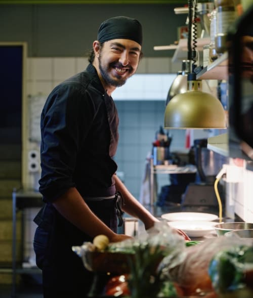 Chef souriant dans la cuisine de l'Hôtel De La Sage avec des ingrédients frais et une lampe chauffante en laiton