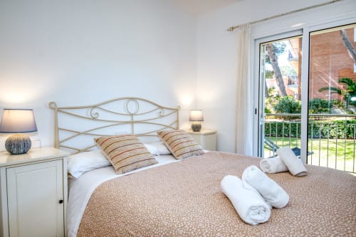 Bright bedroom with ornate metal bed, striped pillows, white bedside tables with lamps, and glass doors opening to garden