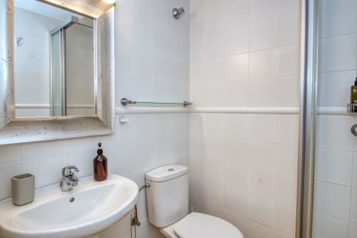 Modern bathroom with white tiles, curved sink, toilet, framed mirror, and glass shower enclosure in apartment at Finques