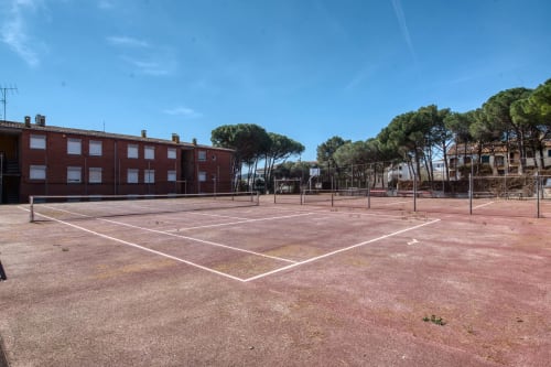 Red clay tennis court with net at Finques Garcia Enrich apartment complex in Palafrugell, Spain, surrounded by pine trees and