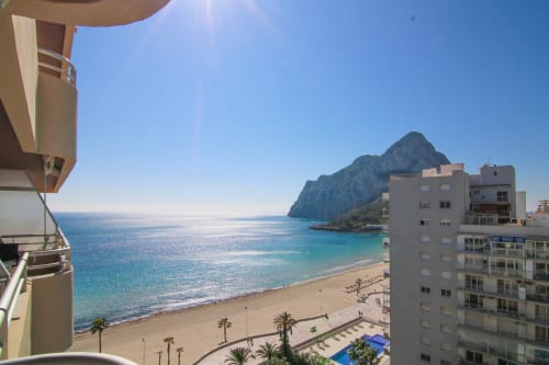 Balcony view of Calpe's Fossa beach, turquoise sea, and dramatic cliff from Riurau Home apartment with palm-lined promenade