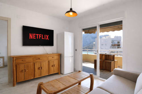 Modern apartment living room with Netflix on flat-screen TV, wooden furniture, balcony overlooking sea in Calpe, Spain