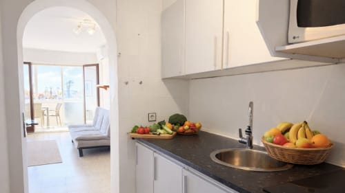 Cocina moderna con encimera oscura, fregadero de acero inoxidable, gabinetes blancos y frutas frescas en canastas