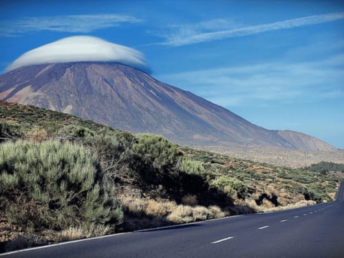 Snow-capped volcanic mountain with lava fields and vegetation along road, Tenerife landscape