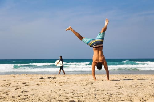 Young man doing handstand on Costa Adeje beach with surfer and turquoise ocean waves in background