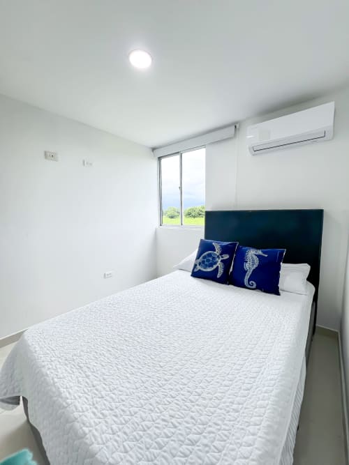 Dormitorio moderno con vista al lago en Olamar Living, Coveñas. Cama blanca, cabecera oscura, almohadas azules, aire acondicionado